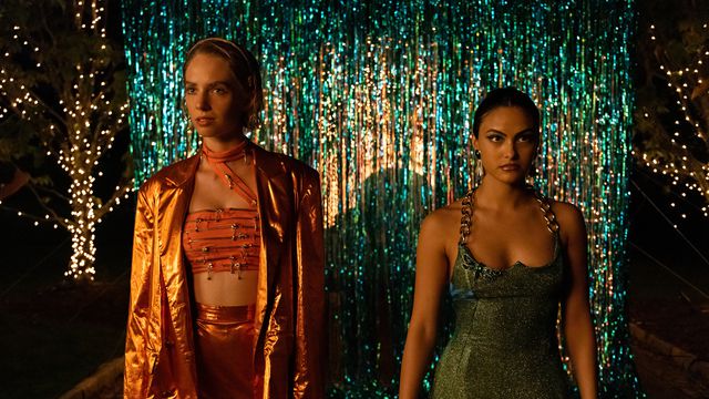 maya hawke dressed in golden outfit next to camila mendes in a green dress. both are in front of a sparkly backdrop