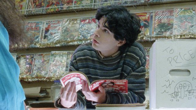Daniel Zolghadri reads a magazine about comic books in a comics store, backed by bagged and shelved comics, in Funny Pages