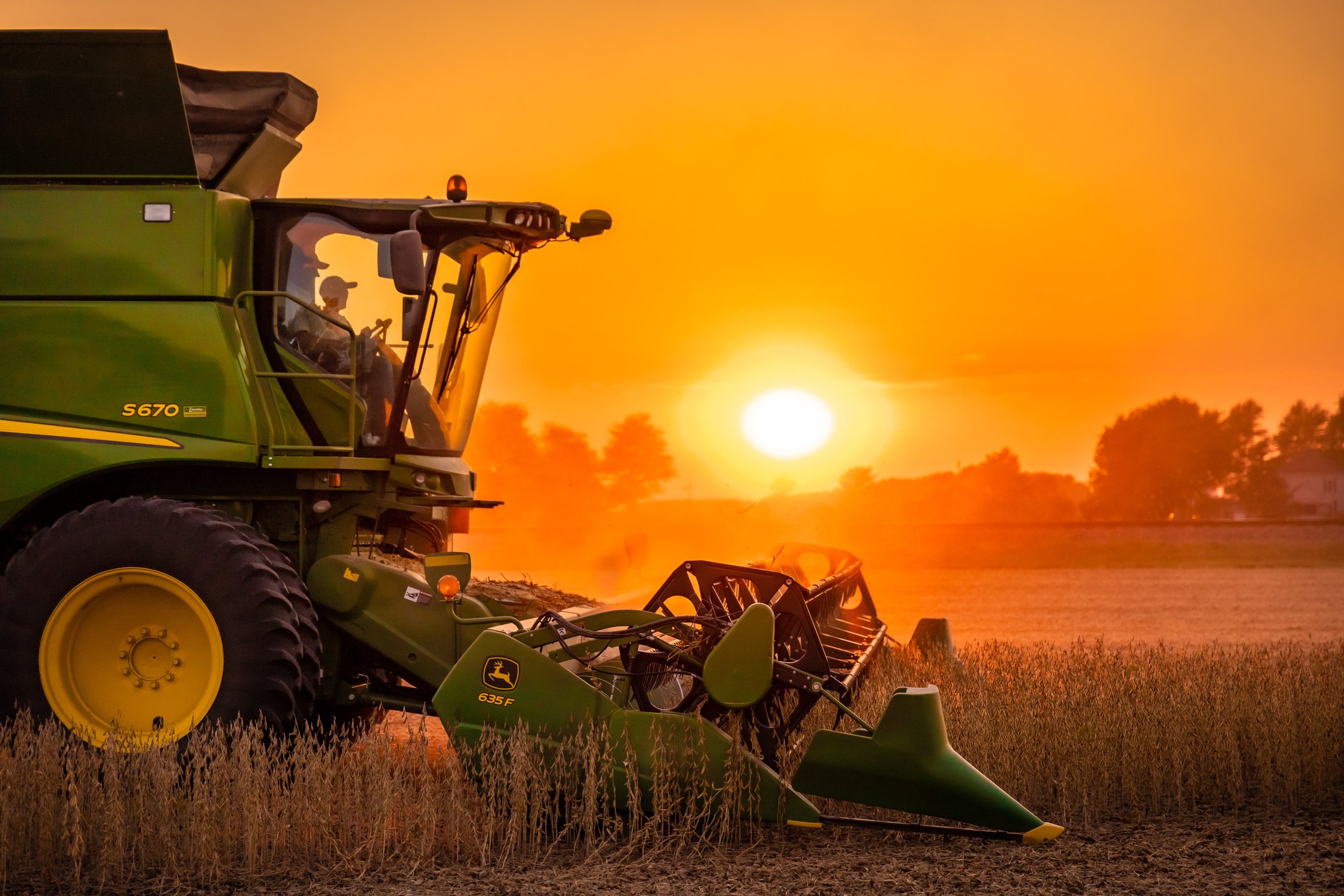 A profile view of a John Deere S670 harvesting soybeans during a hazy sunset with a red and orange sky from the bean dust.