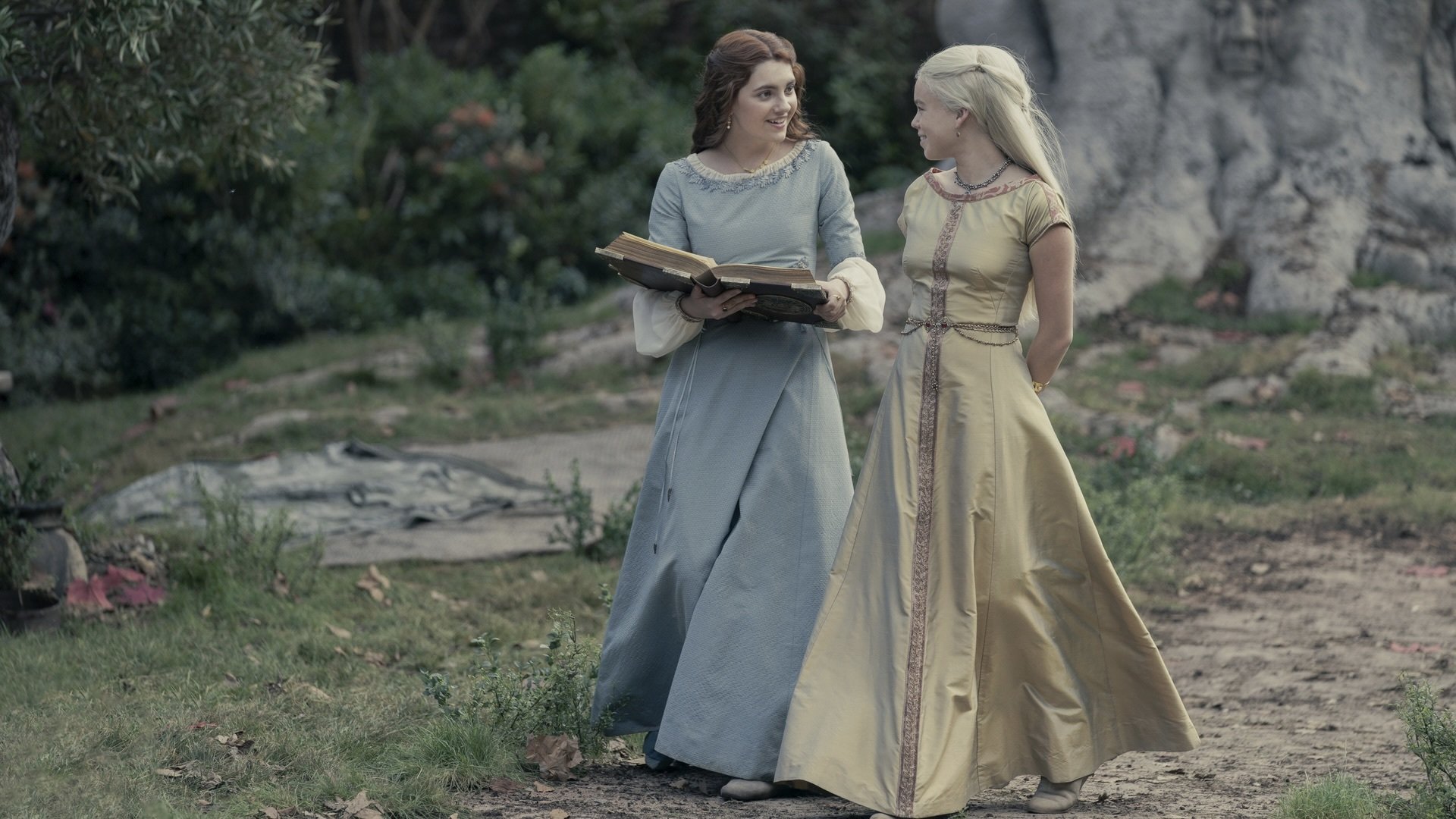 Two young women in lavish fantasy dresses walking through a garden. One holds a large, open book.