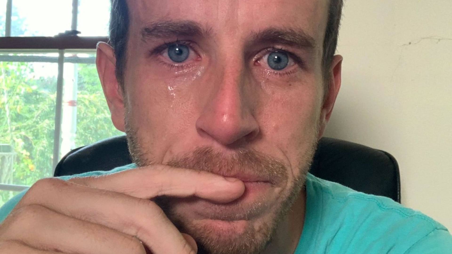 A teary-eyed seflie of Wallake in a bright blue shirt. He has blue eyes and is holding his pointer finger to his lips, which are pursed with emotion.