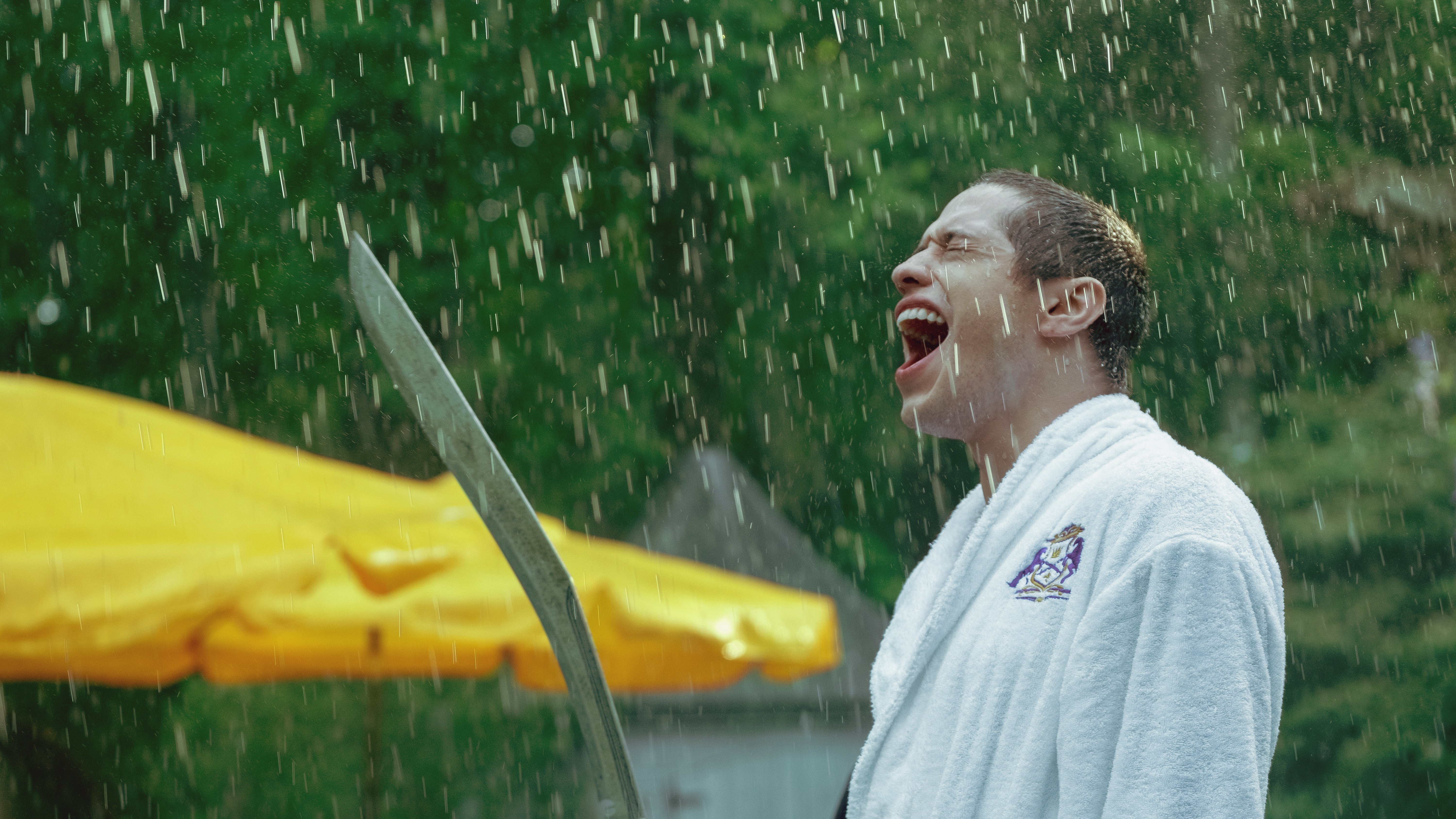 A man wearing a bathrobe and holding a sword stands outside screaming into the rain.