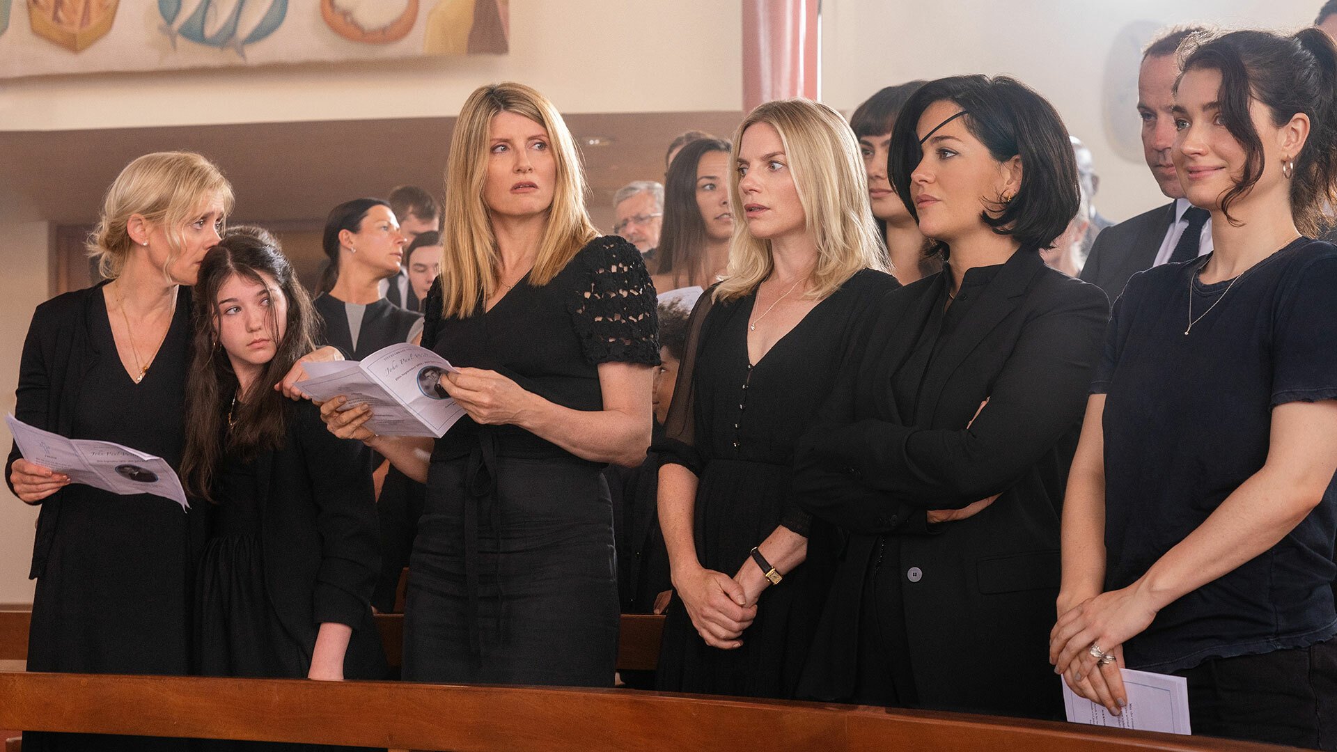 A group of five women stand in the front row of a church dressed all in black, attending a funeral.