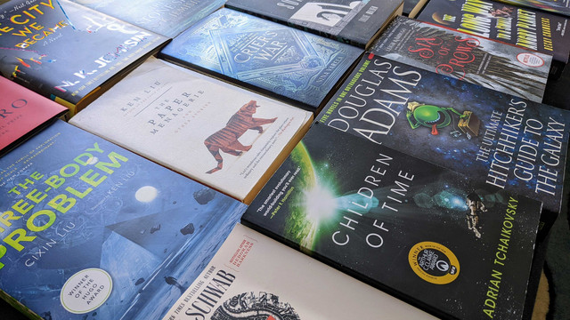 An image of science fiction and fantasy books laid down side by side, shot at an angle. Books include Children of Time, Three-Body Problem, On a Sunbeam, Six of Crows, and others.