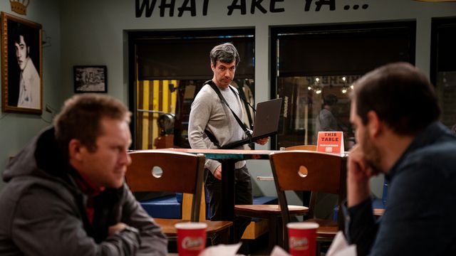 Nathan Fielder standing with a laptop on a sling looking intently at two men chatting over a table in The Rehearsal