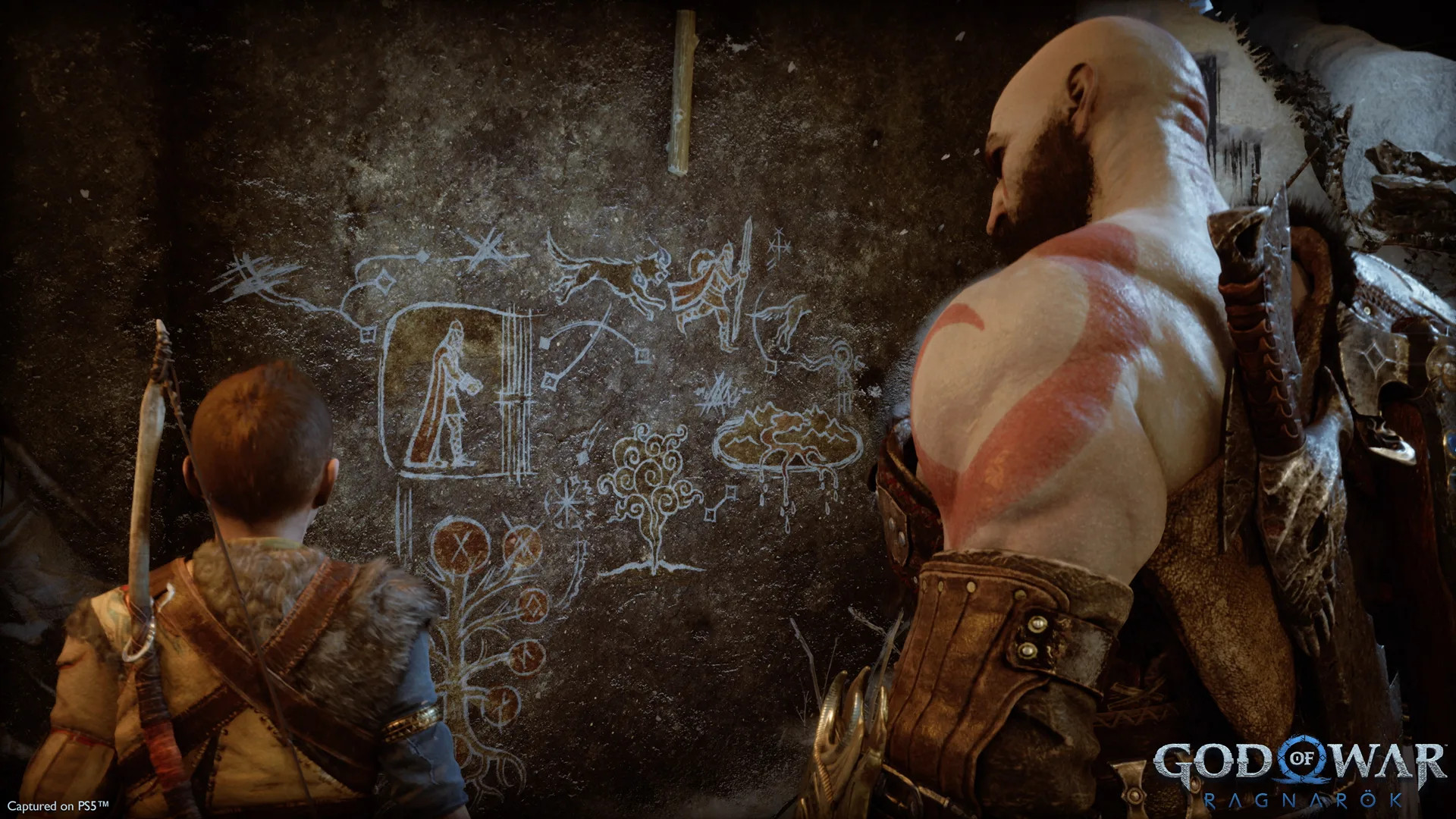 Screenshot from God of War Ragnarok featuring a large man covered in white ash and a red tattoo and his son, a young boy clad in furs and carrying a boy pondering cave drawings