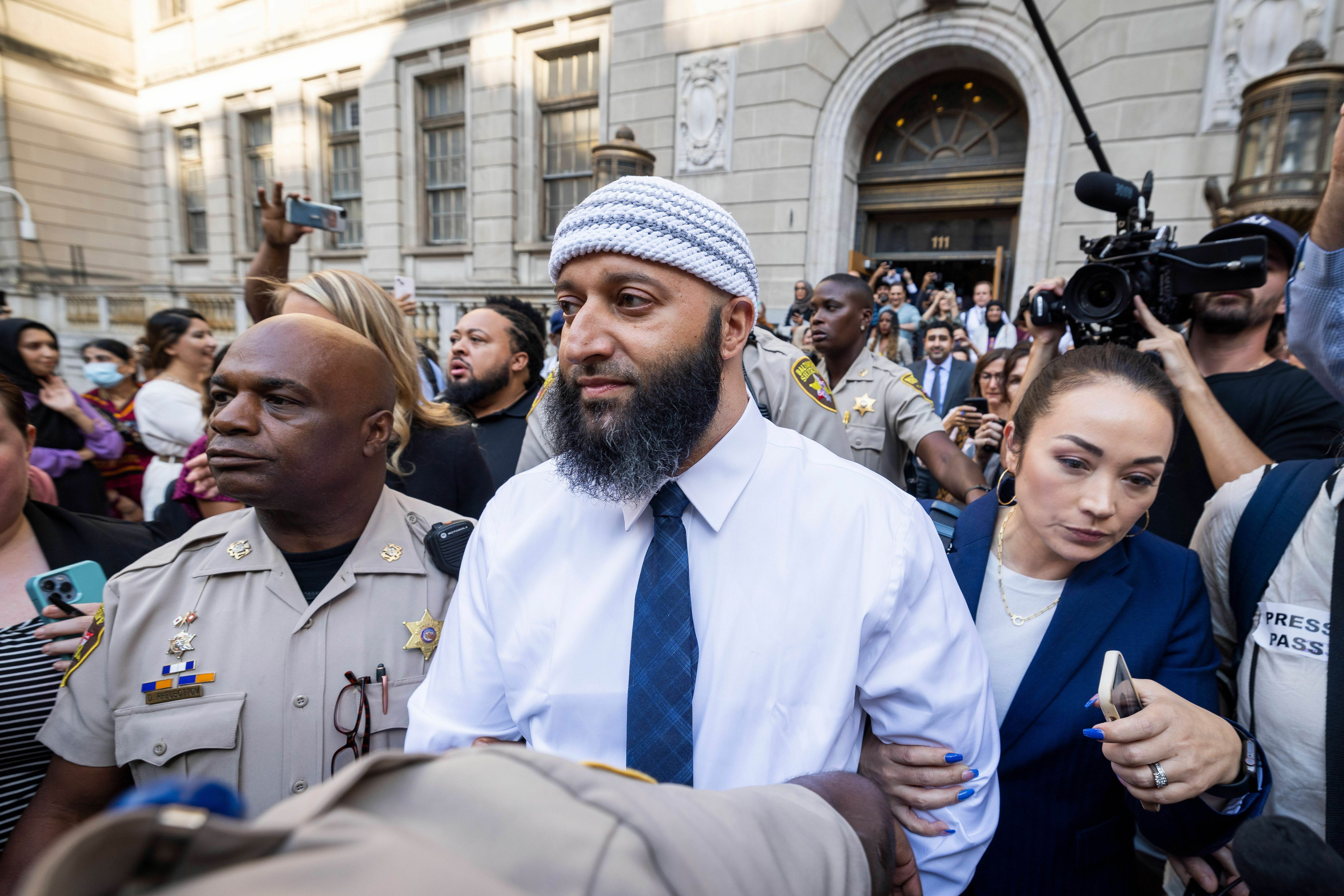 Adnan Syed walks down the steps of the courthouse, surrounded by press microphones as his lawyer stands next to him.