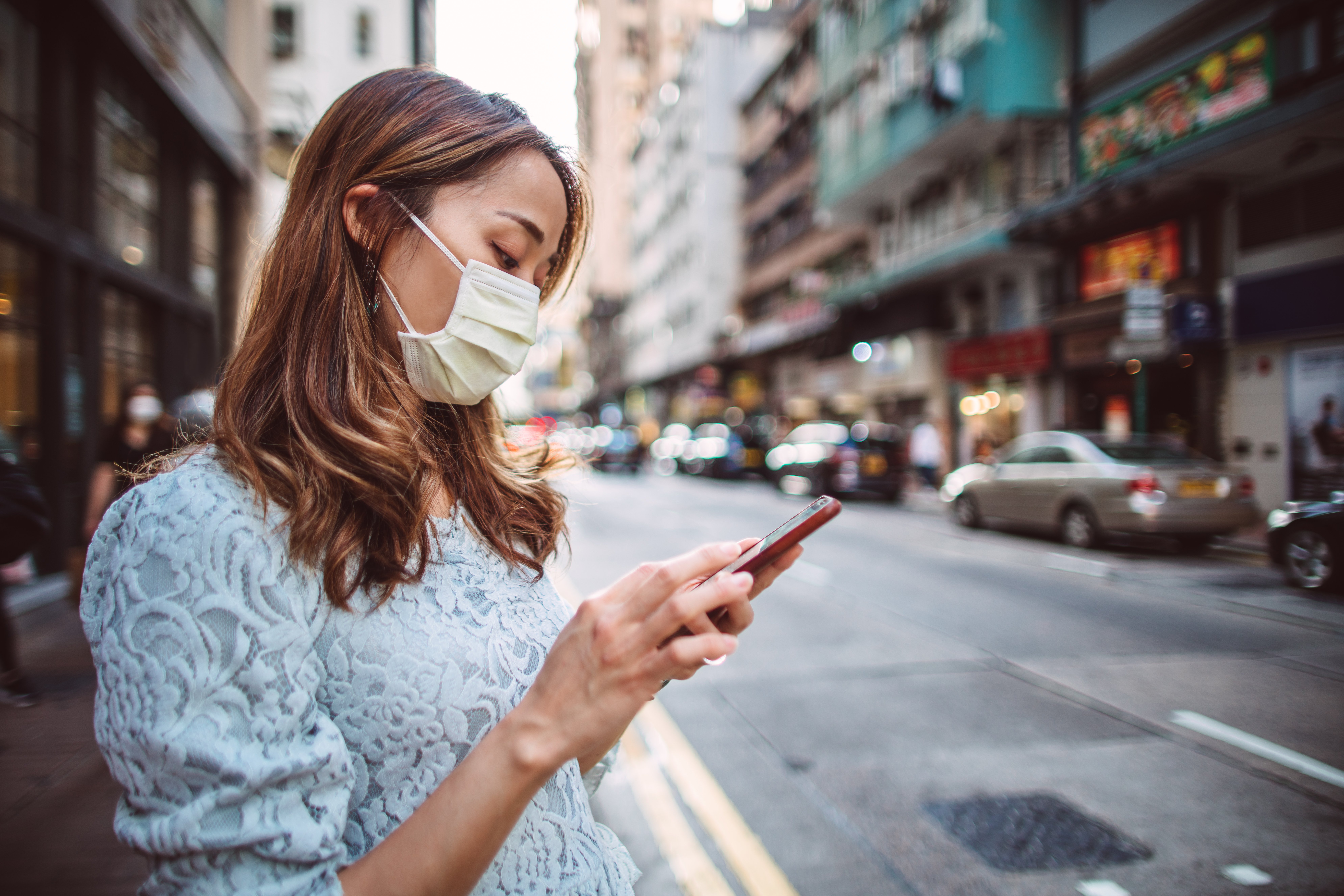 A woman in a mask users her smartphone next to a city street