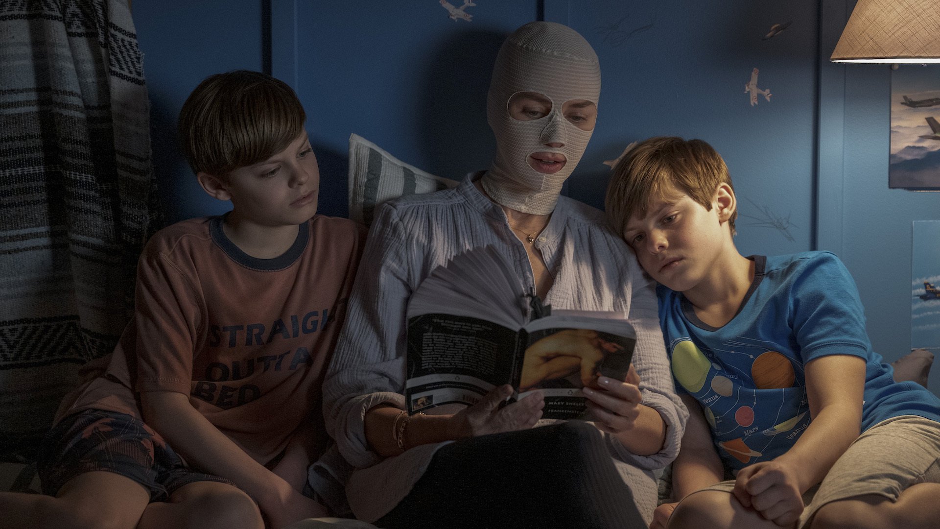 A woman in face bandages reads to twin boys in bed.