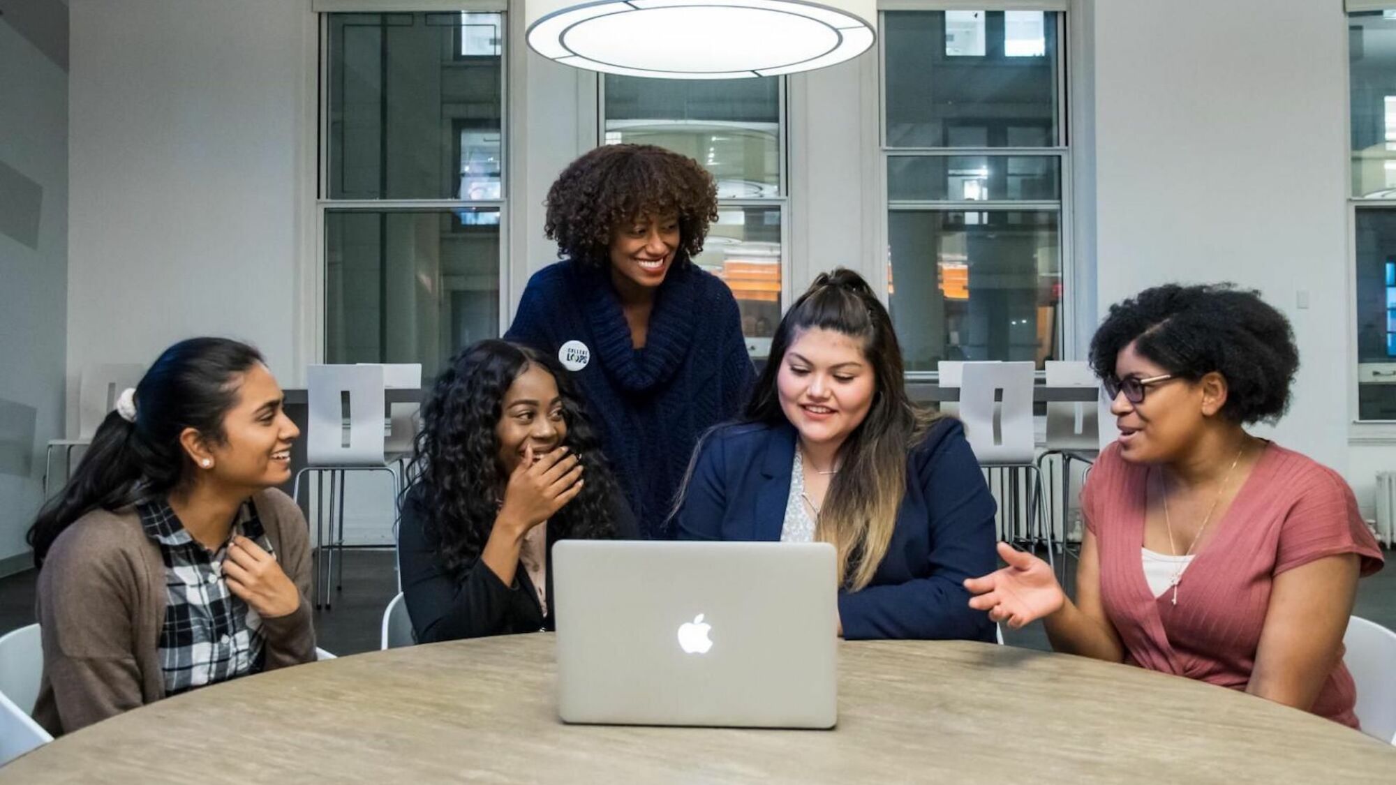 Five women stand around a laptop in an office smiling.