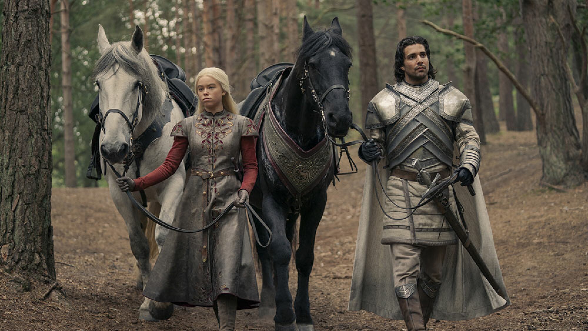 A blonde woman and a knight walk through the woods with their two horses.