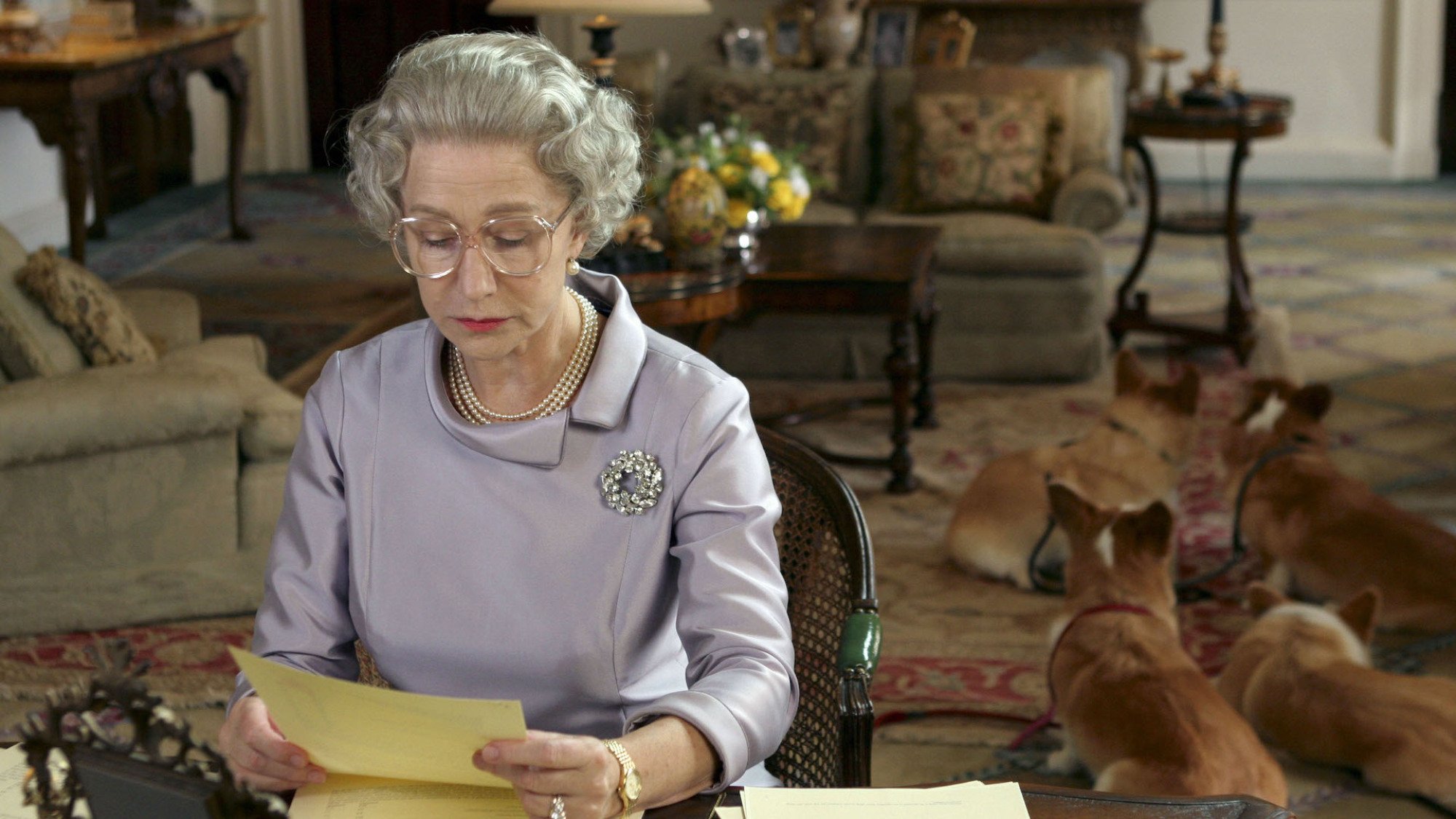 An actor dressed up as Queen Elizabeth II reading letters with her corgis in the background.