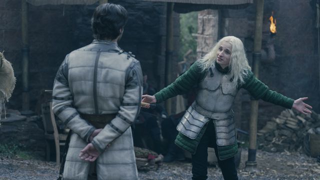 Ty Tennant from HBO’s House of the Dragon as Aegon Targaryen, spreading his arms out wide as a challenge to Ser Criston Cole. He wears plate armor and long green sleeves, and has bright long hair.