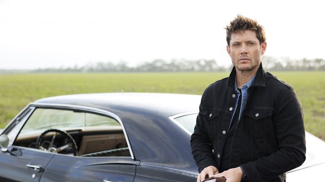 Jensen Ackles as Dean standing by his black camero car next to a green field in The Winchesters