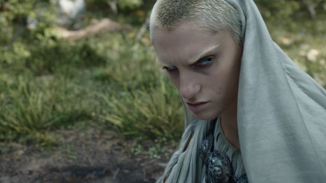 Bridie Sisson, a blond-haired actor, with a gray shawl draped over the back of her head, looks suspicious in The Lord of the Rings: The Rings of Power.