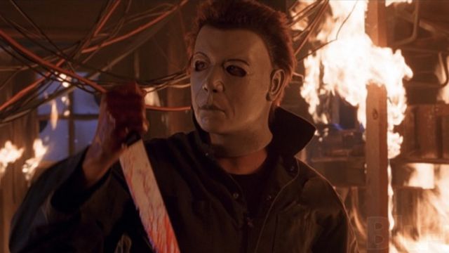 Michael Myers, surrounded by flames, wears a mask with arched eyebrows and a more distinct nose. He holds a bloody knife.