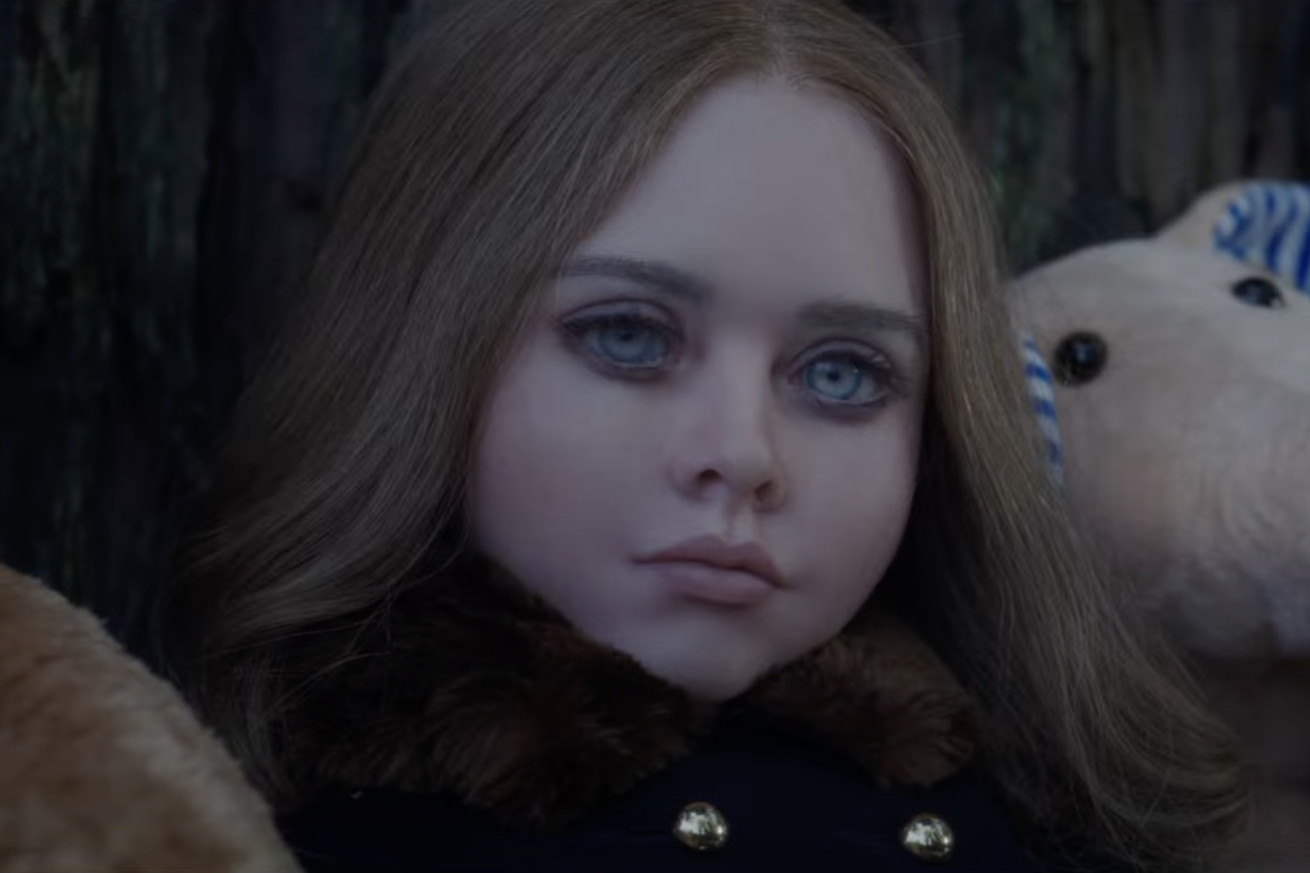 A tight shot of a very lifelike but decidedly off-putting and unsettling doll made to look like a young girl with abnormally large eyes that are piercing blue. The doll is making a visibly judgmental face and is flanked on either side by regular stuffed animals.