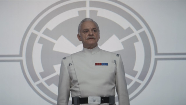 Colonel Wullf Yularen (Malcolm Sinclair) standing in his white military uniform adorned with red and blue rank pins, giving a speech in the seventh episode of Andor season 1