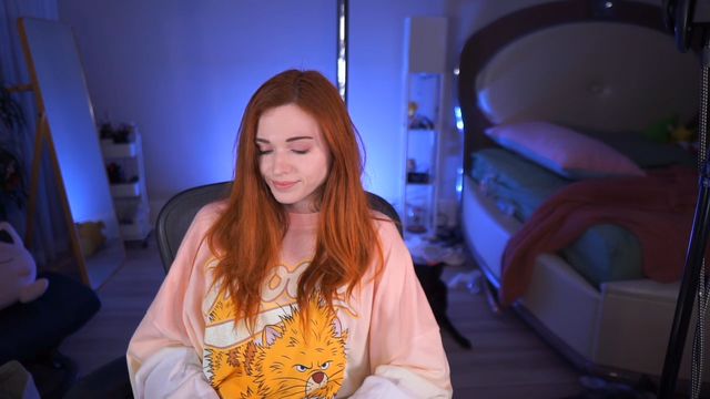 Twitch streamer Amouranth, a light-skinned woman with long red hair, is sitting and wearing a large sweatshirt. She is smiling and looking down.