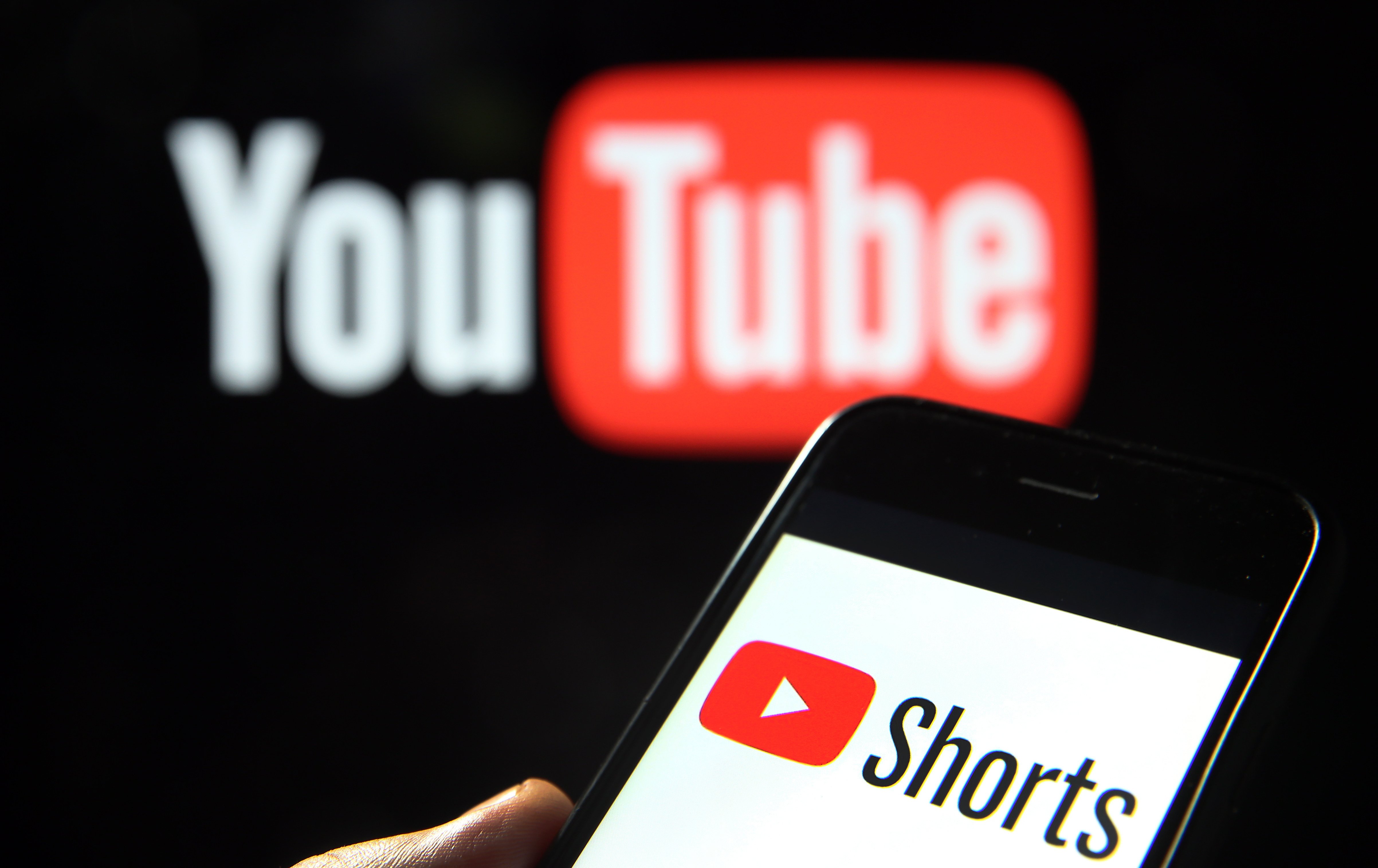In the foreground, a hand holds a phone with the YouTube Shorts logo visible in red, white and black. In the background and out of focus is the YouTube logo.