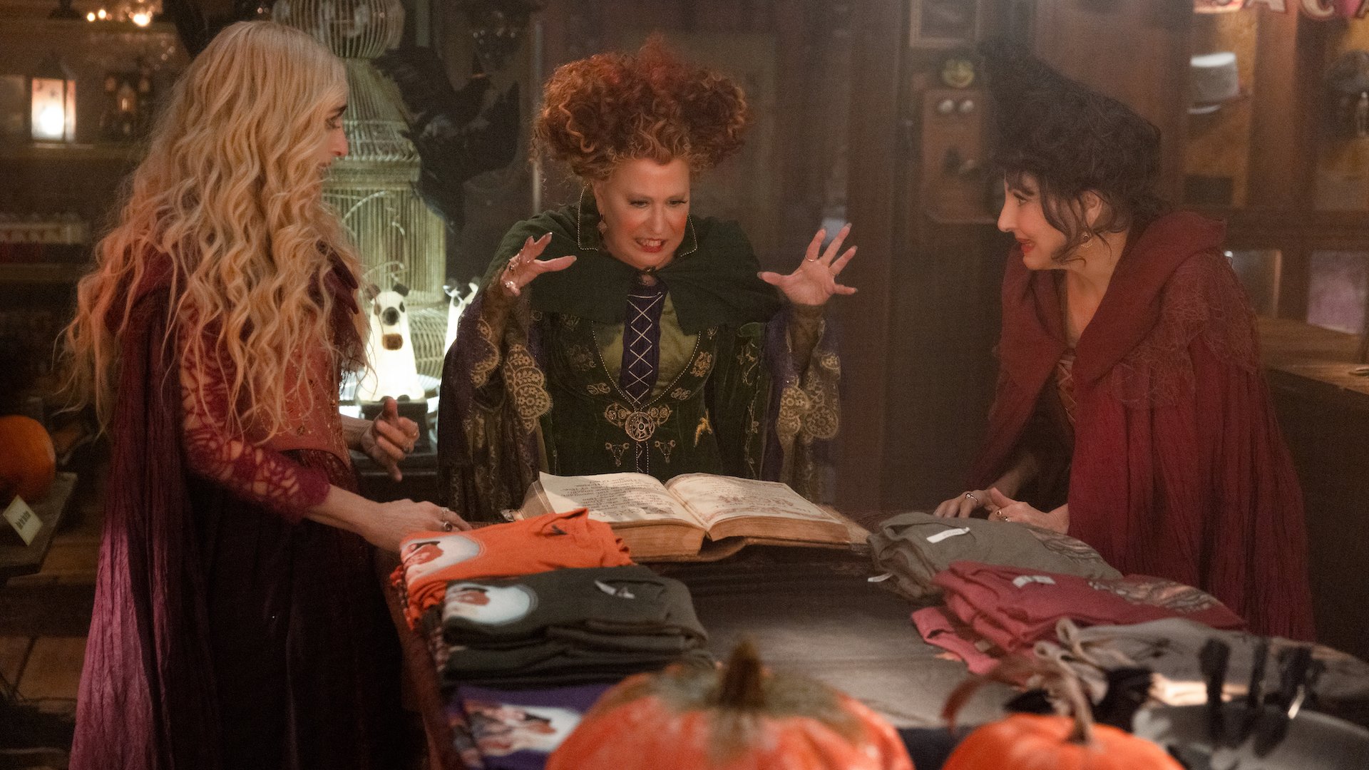 Three witches stand around a spell book and table laden with spell things.