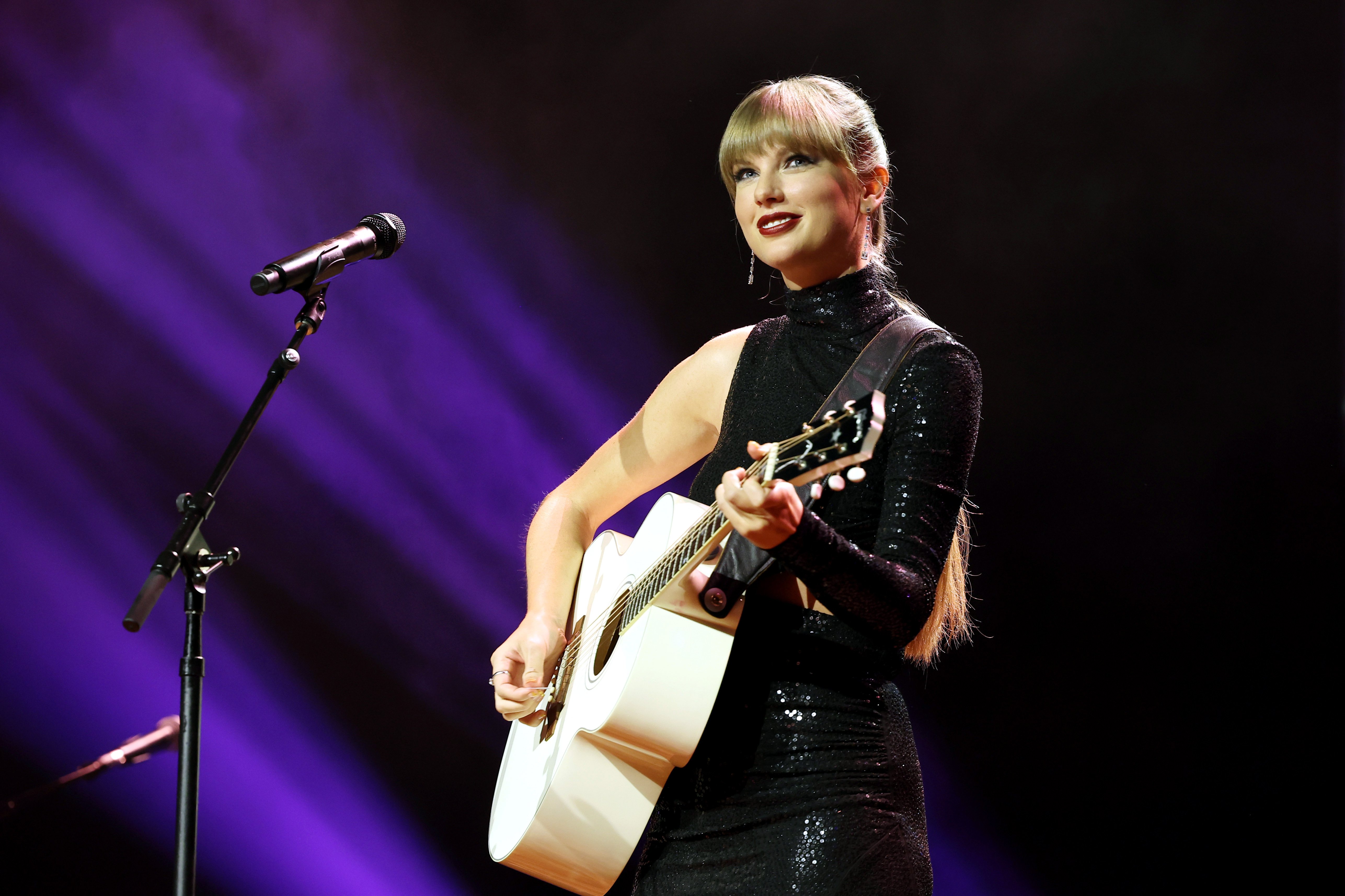 Taylor Swift, in a black single-sleeved dress, stands in front of a microphone while holding an acoustic guitar. 