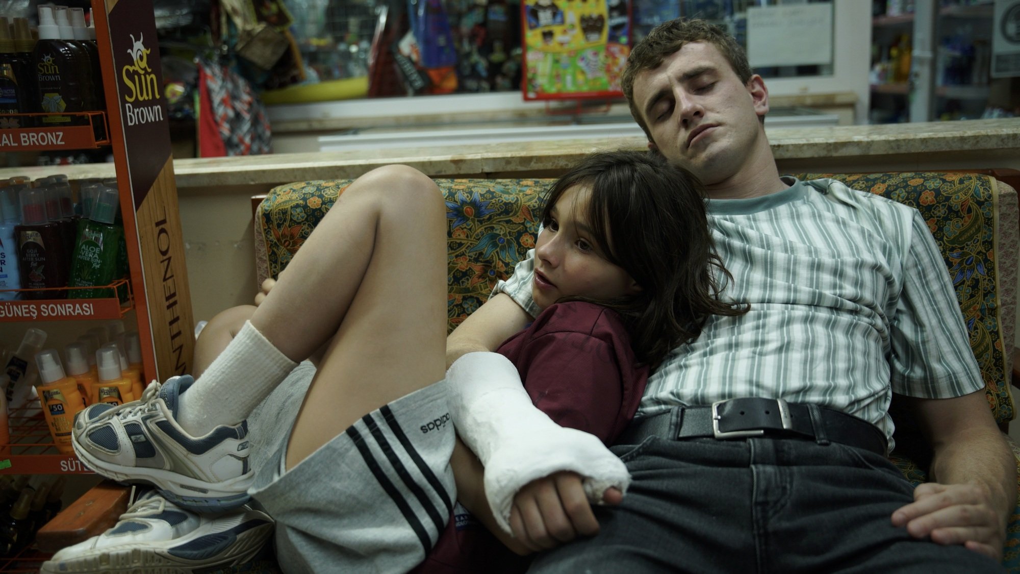 A man in an arm cast sits on a couch with his young daughter, wrapping his arm around her.