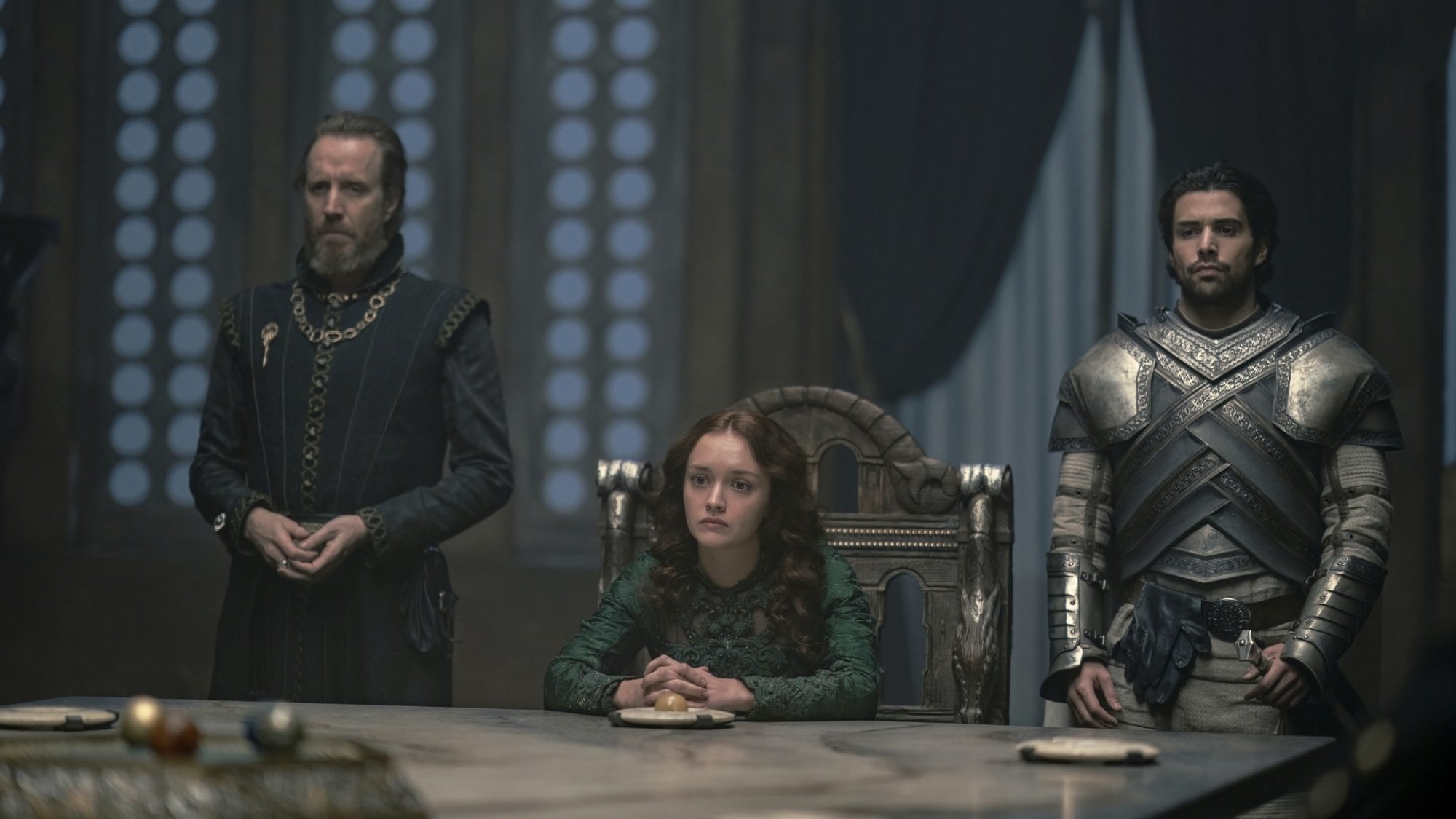 A woman in a green dress sits at the head of a large table. A man in knight's armor stands on one side of her, and a man in black clothing with a bronze hand pin stands on the other side.