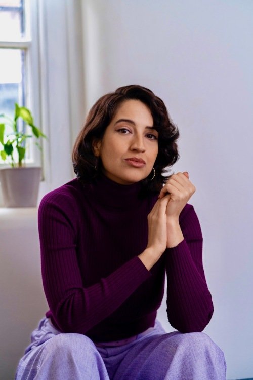 A woman in a maroon turtleneck sweater sits in a light room with a plant in the background.