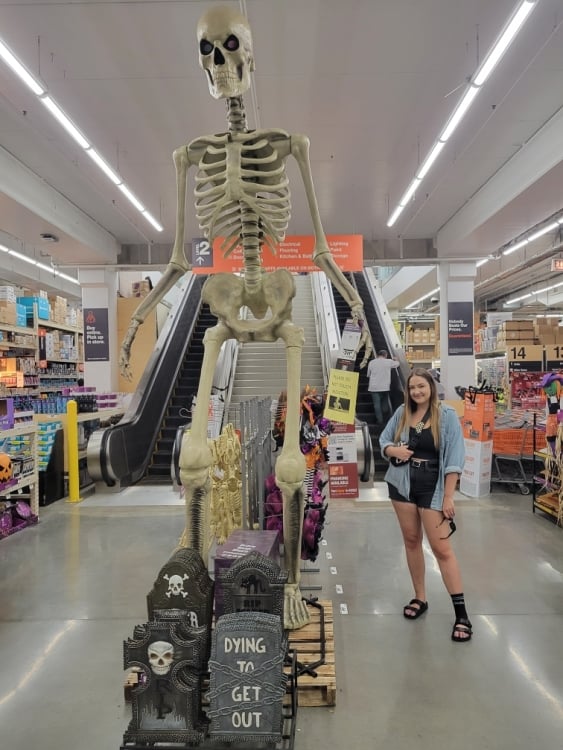 a woman standing next to a 12-foot skeleton in the middle of a home depot