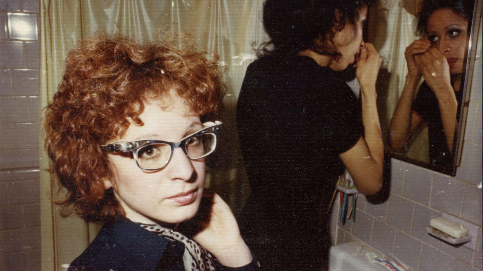 Two women in a bathroom: one of them, with red curly hair and glasses, looks directly in the camera, while the other applies makeup in the mirror.