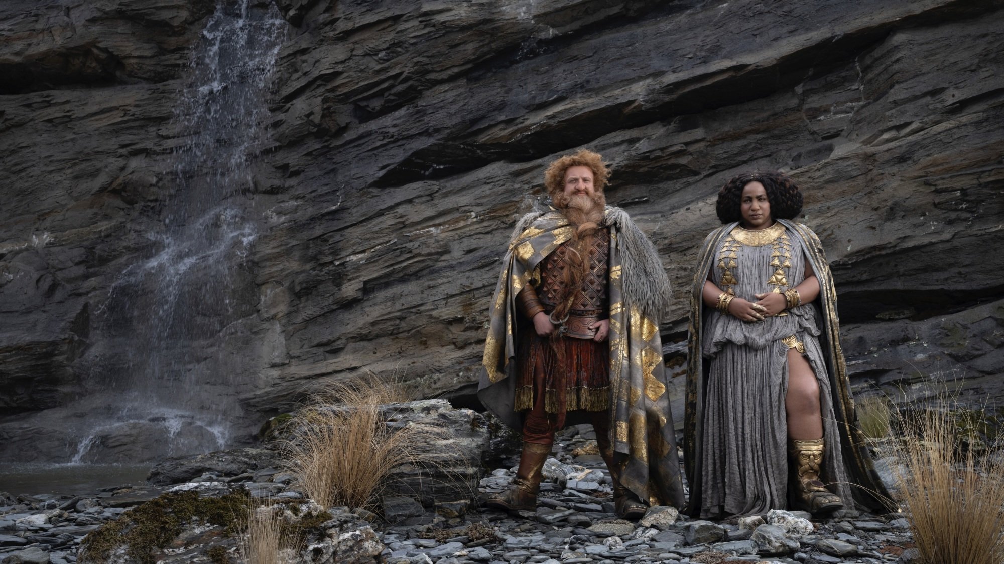 A dwarven man and woman in lavish fantasy clothing and armor stand in front of a rock face with a waterfall falling down it.
