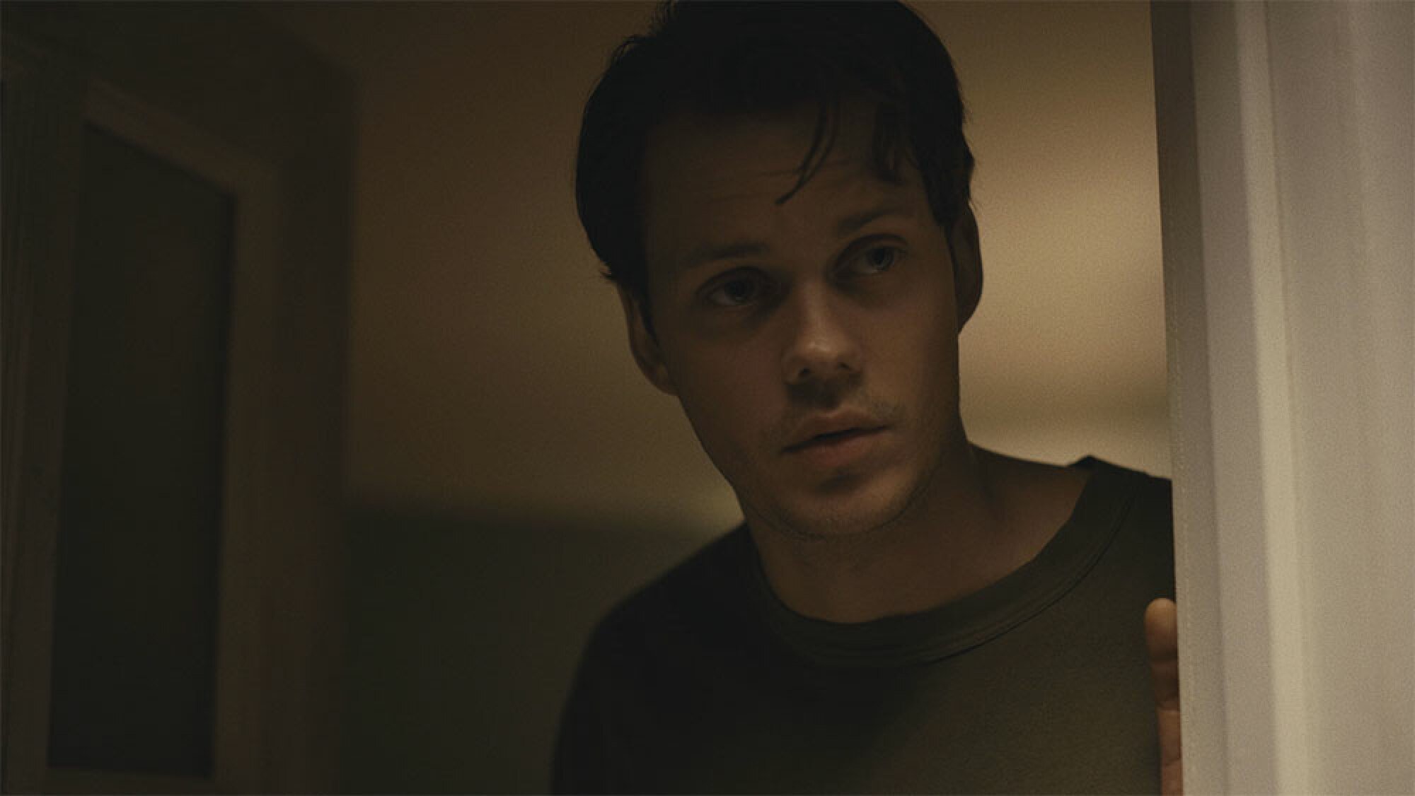 A dark-haired man stands in an open front door, peering out into the night.