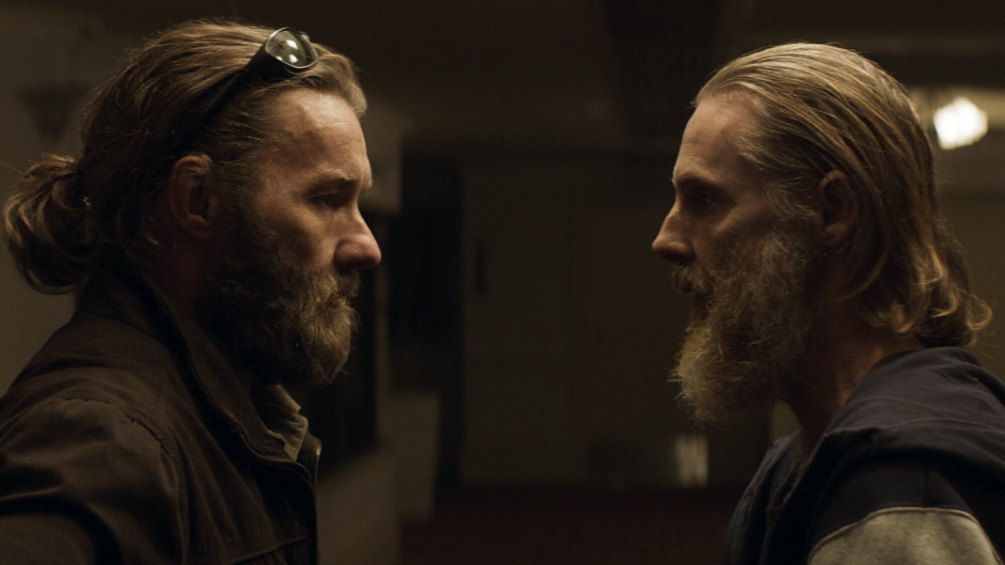 Two bearded men stand in a room staring at each.