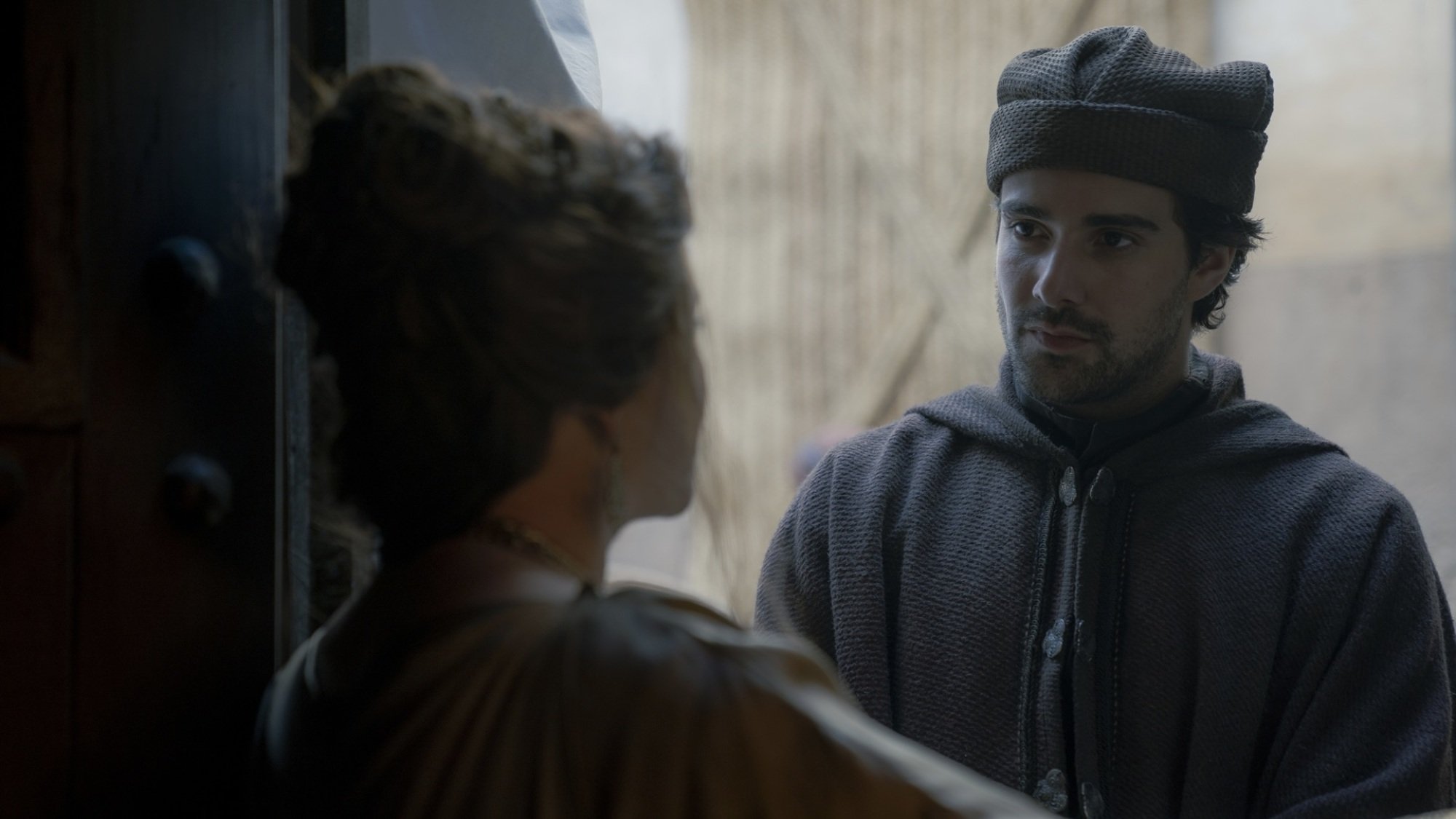 A man in a brown cloak and hat stands in a door, listening to a woman speak to him.