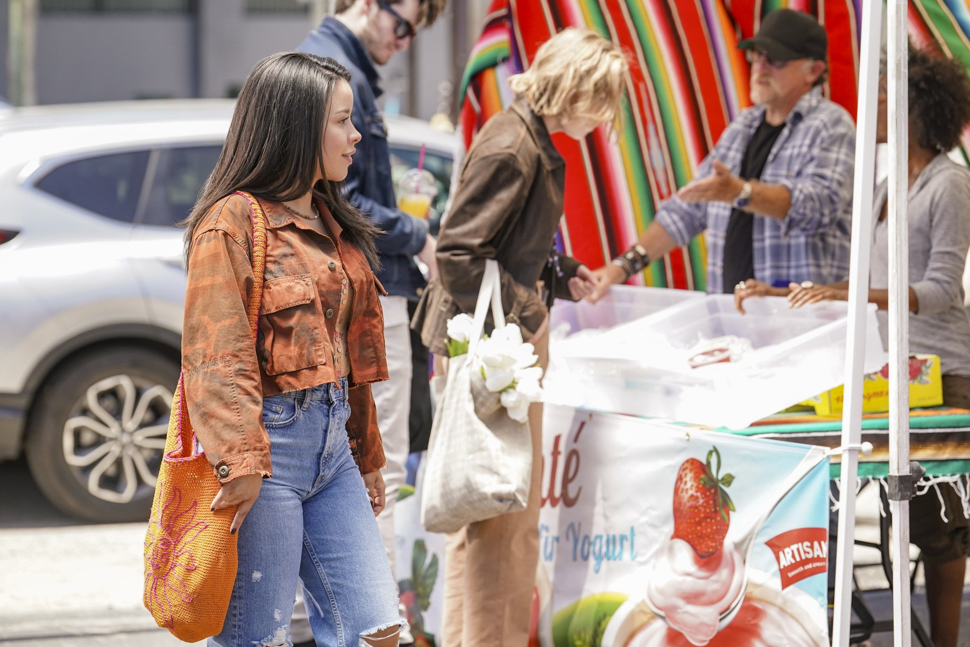 A young woman wearing an orange jacket and bag walks past a farmers' market stall.
