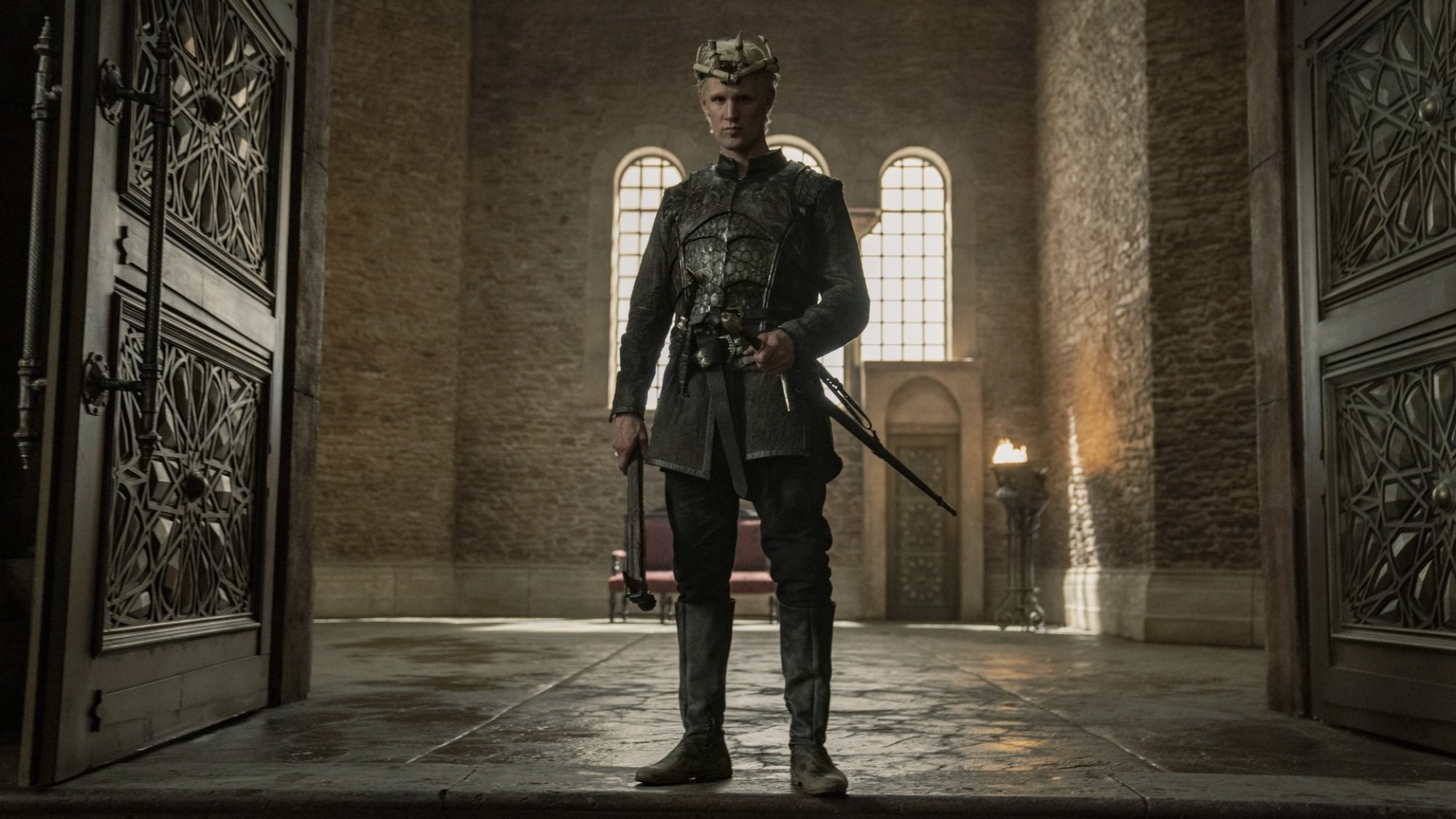 An armored man with silver blonde hair and a crown made of driftwood stands in the door to a great hall.