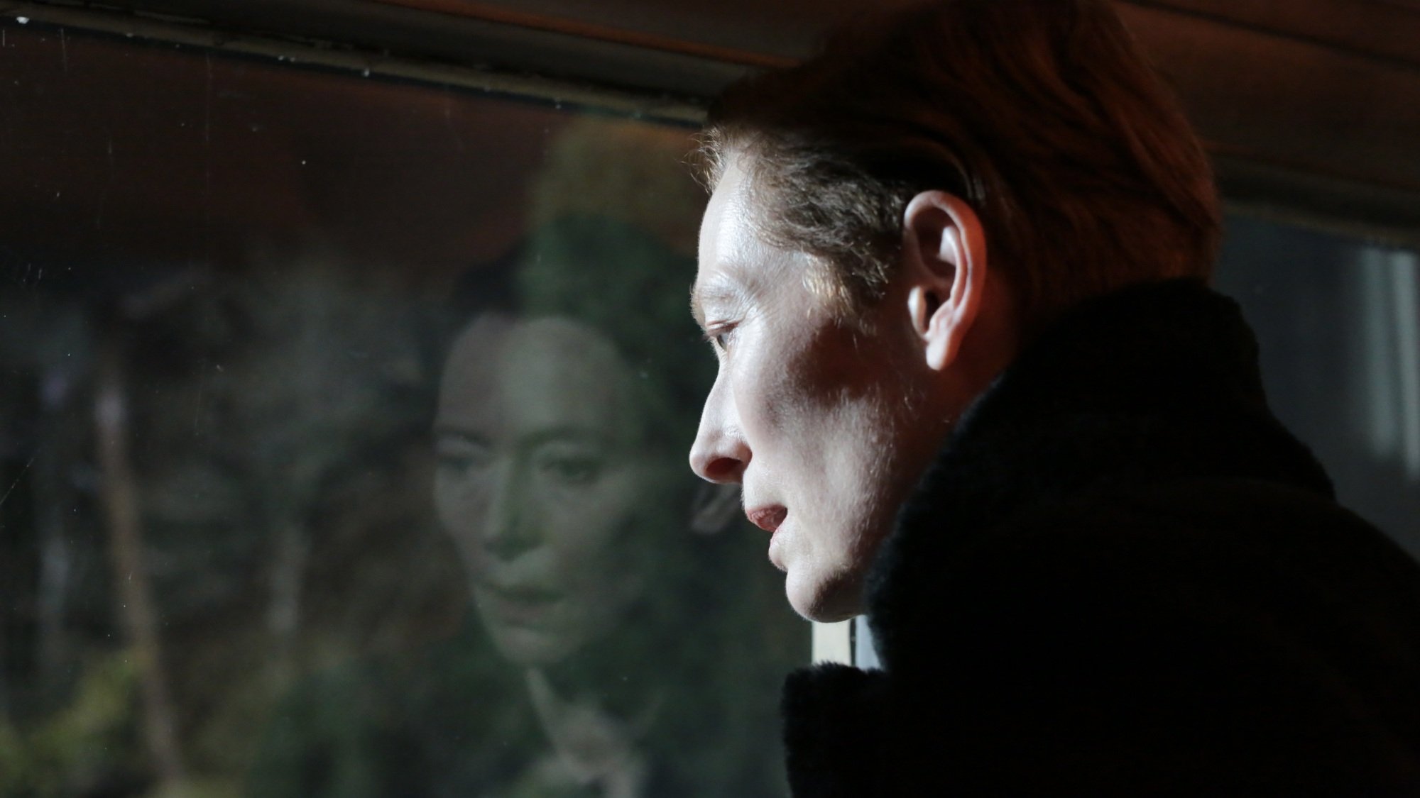 A woman looks out a window — her face is reflected in the glass.