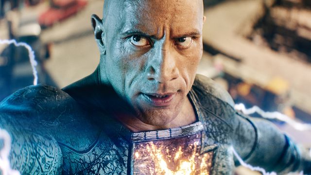 Black Adam (Dwayne Johnson) stands in a battle pose as electricity emanates from his body and the lighting bolt symbol on his black suit lights up in Black Adam. 