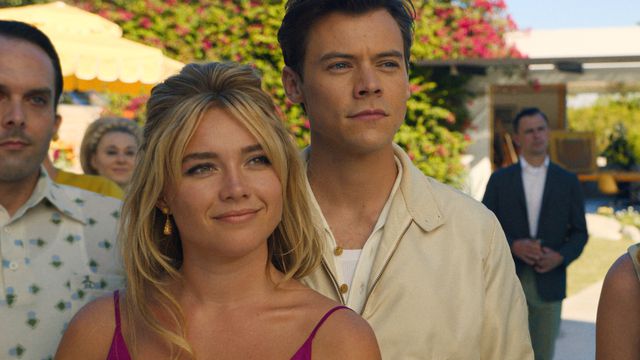 Florence Pugh as Alice and Harry Styles as Jack smile at a sunny garden party, surrounded by other characters, in Don’t Worry Darling.