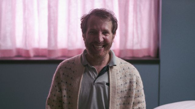 A redheaded bearded man in a sweater sits on a hospital bed in front of pink curtains with the biggest smile ever