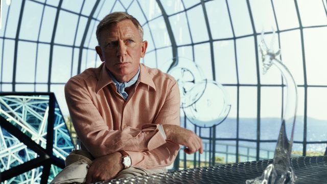 Daniel Craig as Benoit Blanc in Glass Onion, looking thoughtful in a pink linen shirt and blue cravat inside a multifaceted glass dome
