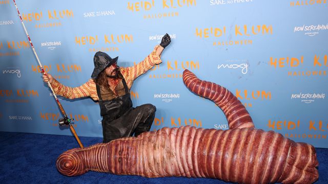 A photograph of the German American model Heidi Klum and her husband Tom Kaulitz dressed in their Halloween costumes at Klum’s Hallowe’en Party 2022. Klum wears an eerily realistic worm costume and is laying down on the ground — No but seriously, it looks super fleshy and gross. 