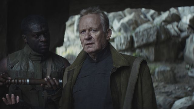 Stellan Skarsgard as Luthen Rael looks forward while standing next to another man in Andor. Rocks are in the background, and the other man holds out an object Luthen is reaching for.