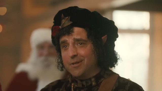 bernard the elf smirking at the camera; he is a middle-aged man with curly dark hair and warm brown eyes, wearing a festive dark green beret