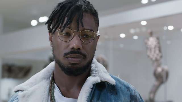 Erik Killmonger (Michael B. Jordan) in a fleece-lined jean jacket and glasses stands in a museum in 2018’s Black Panther