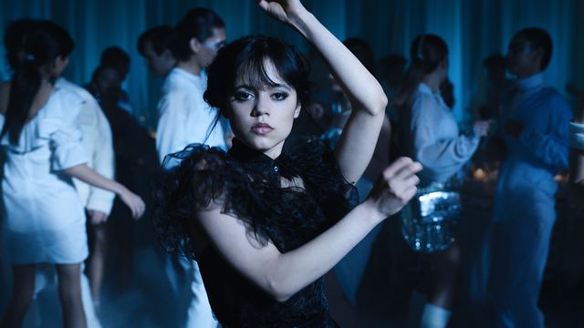 wednesday wears black in a sea of white dresses; she dances chaotically, one arm over her head and the other swung in front of her body; her face is remarkably serious