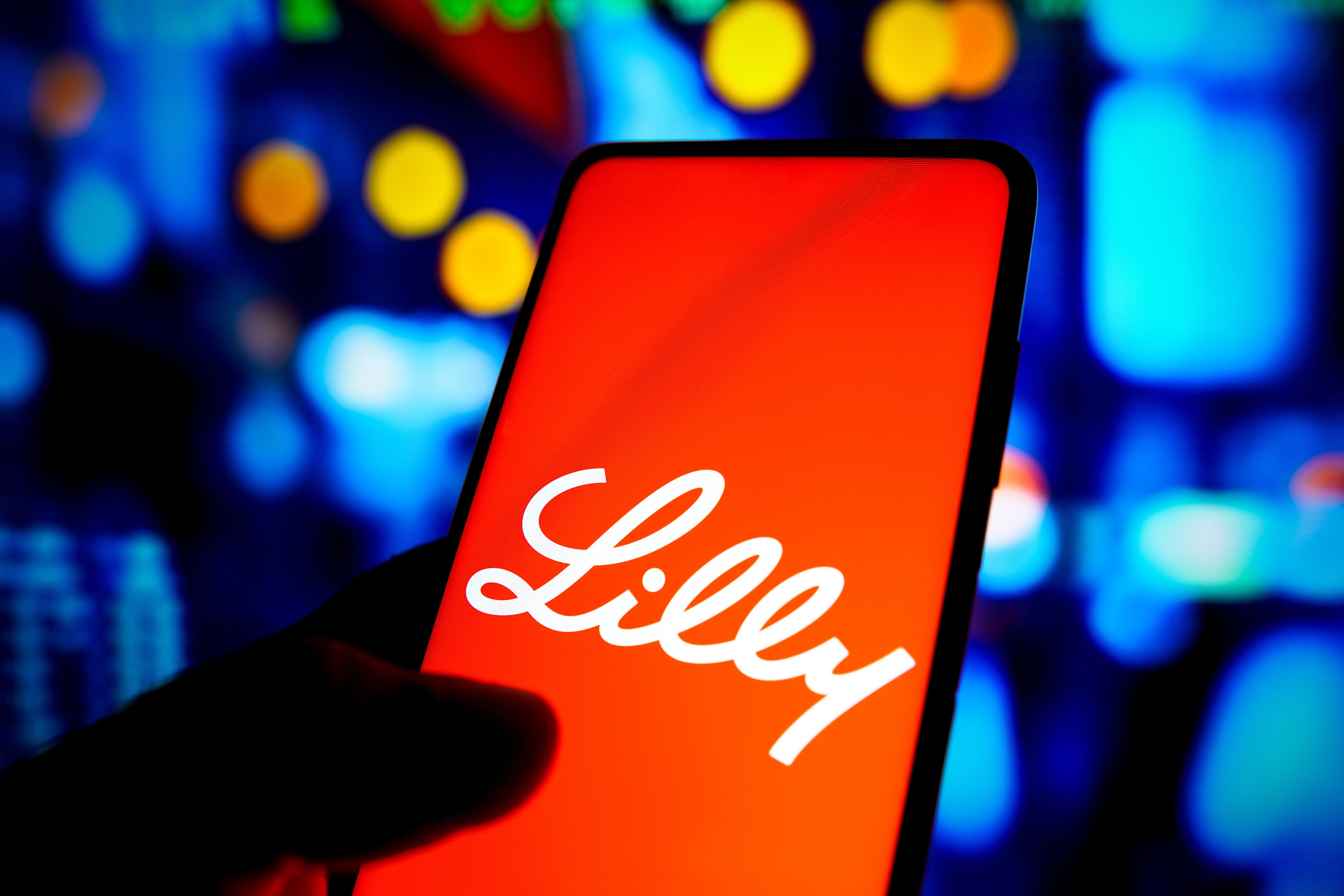 A hand holds an iPhone in front of a blurred multicolored background. The iPhone screen displays the Eli Lilly company logo.