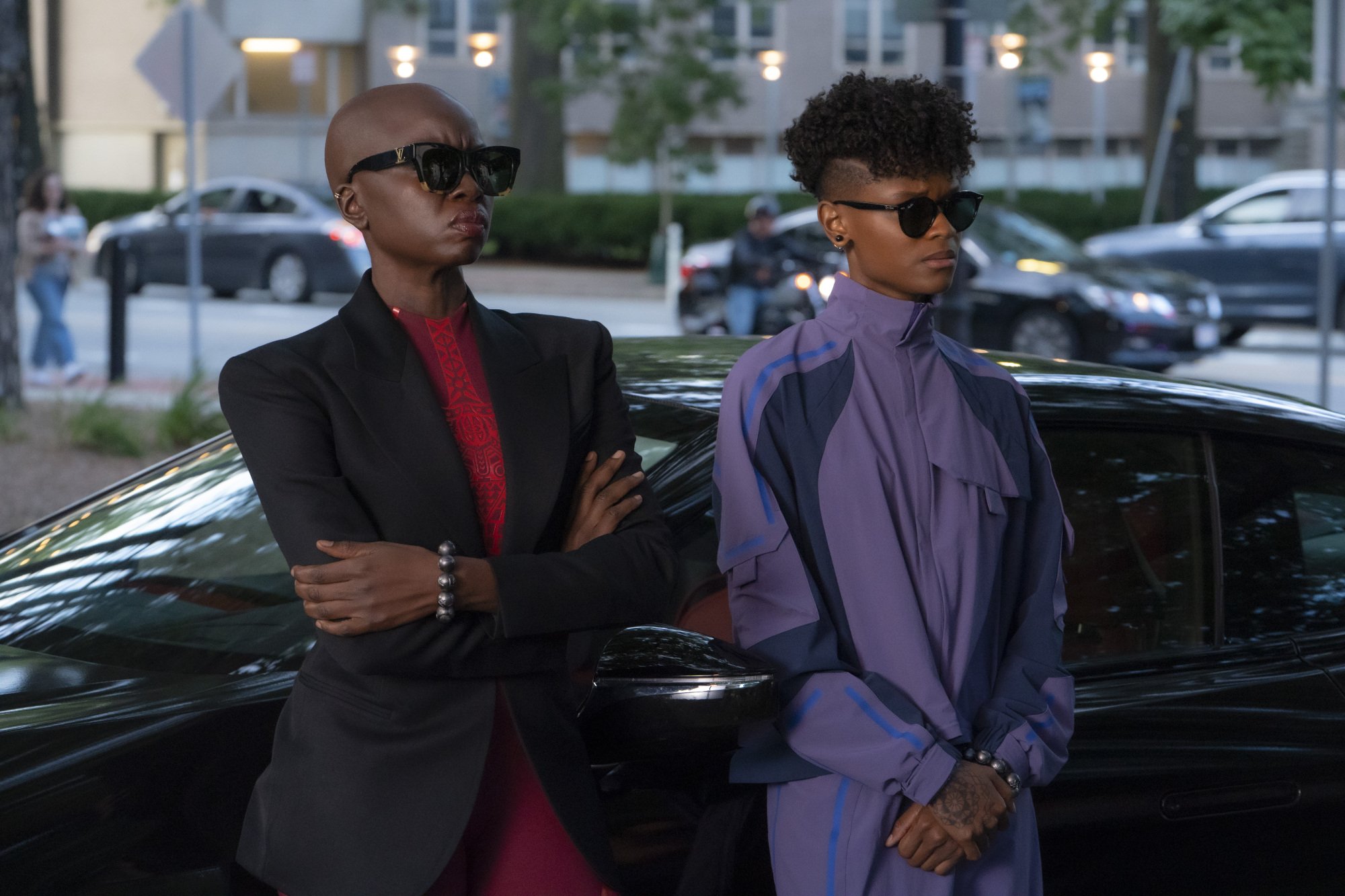 Two Black women in sharp outfits and sunglasses stand next to a sleek car.