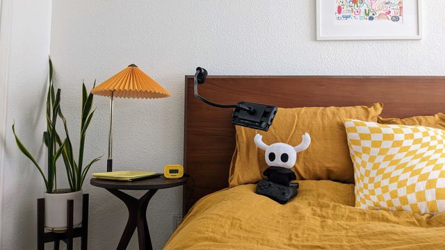 A Hollow Knight plush sits on a bed in front of a mounted Nintendo Switch, with a Switch pro controller in front of it. It is on a bed with yellow sheets.